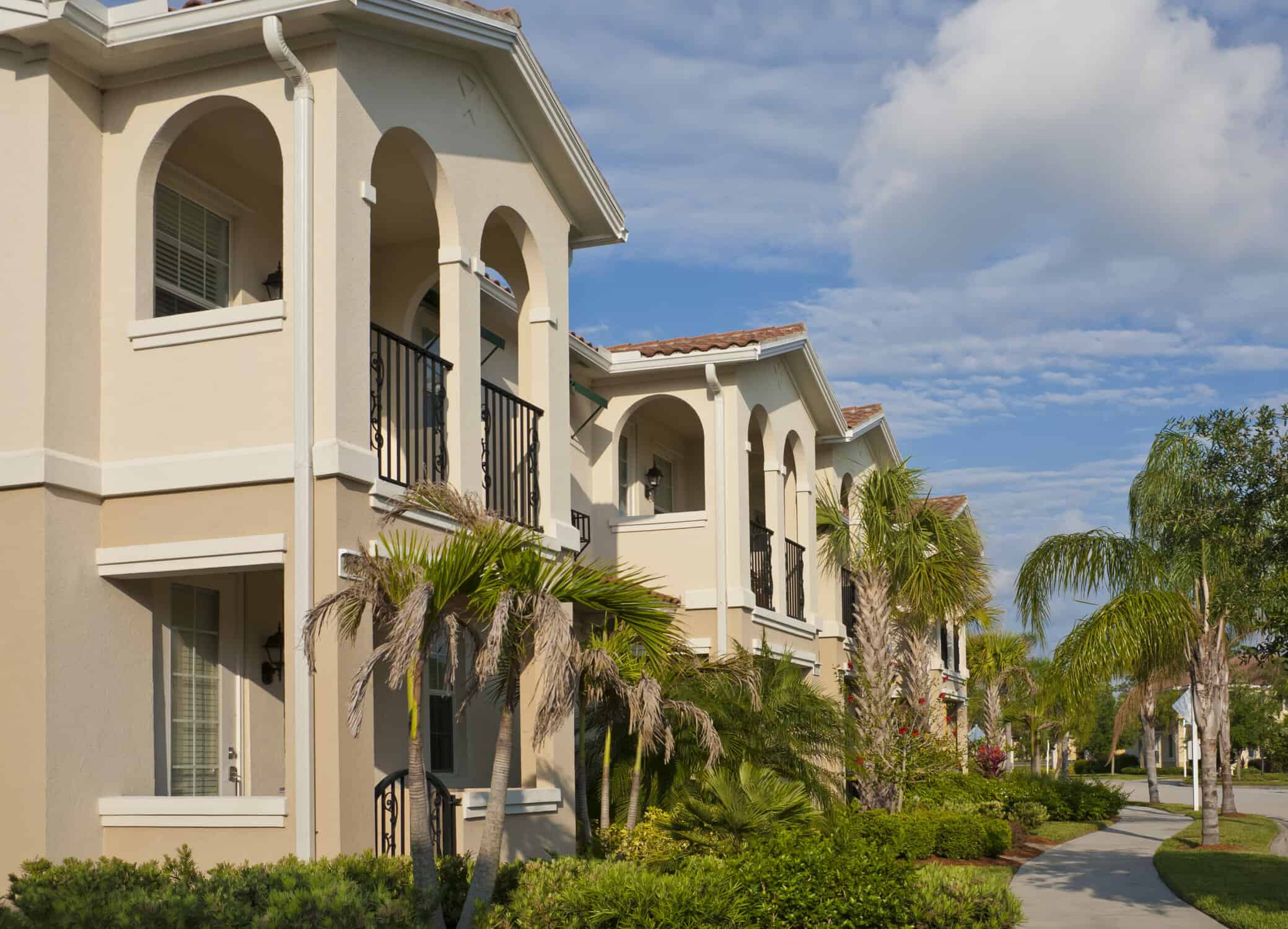 A row of two-story condominiums with sidewalks, palm trees, and tropical foliage.